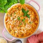 Overhead view of a pink Dutch oven of ground pork pasta topped with parmesan and basil leaves.