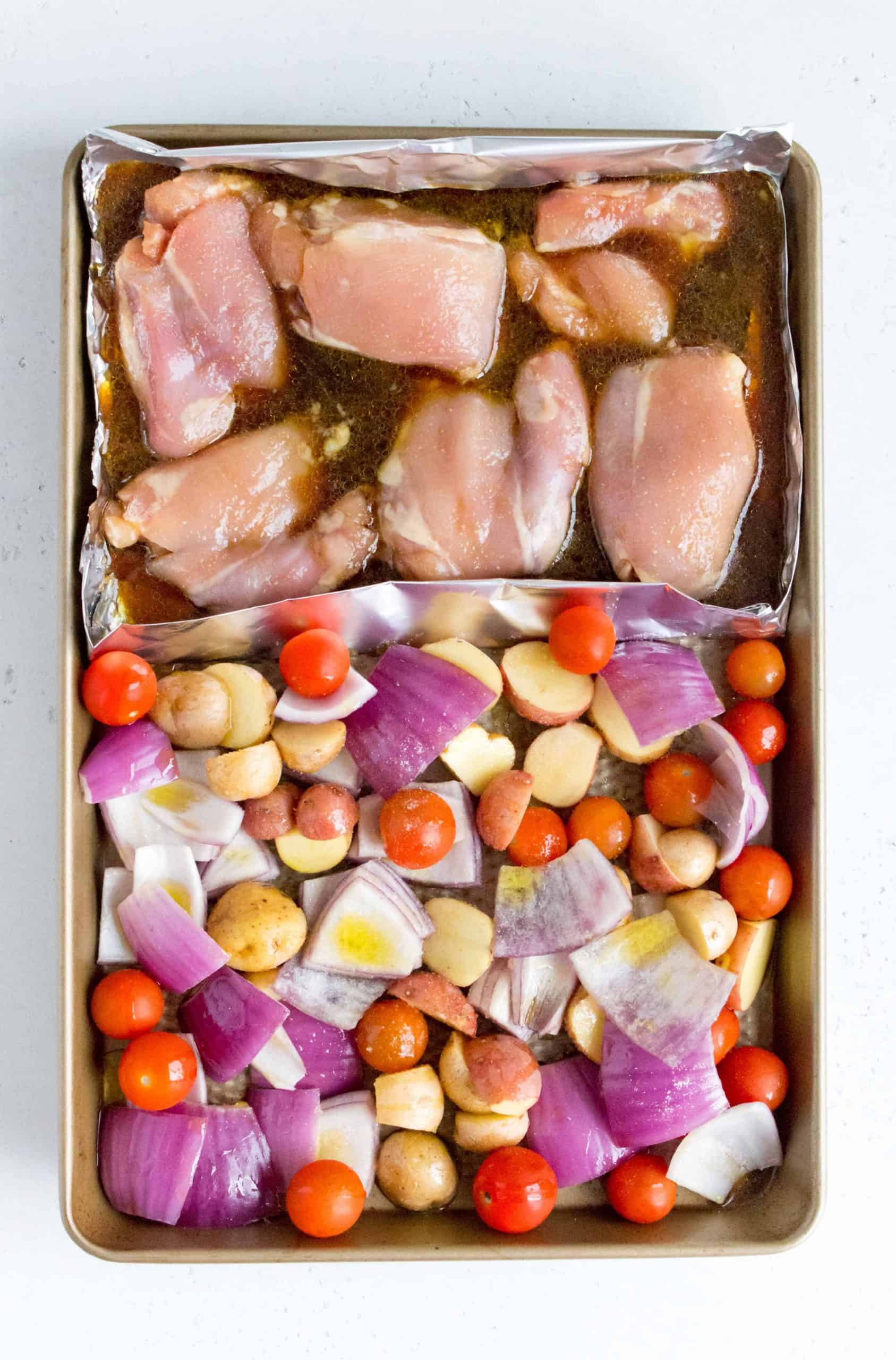 Overhead view of a sheet pan with honey soy chicken with potatoes, red onions, and grape tomatoes before roasting.