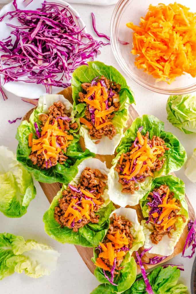 Multiple beef lettuce wraps on a cutting board with lettuce, cabbage, and carrots around them.