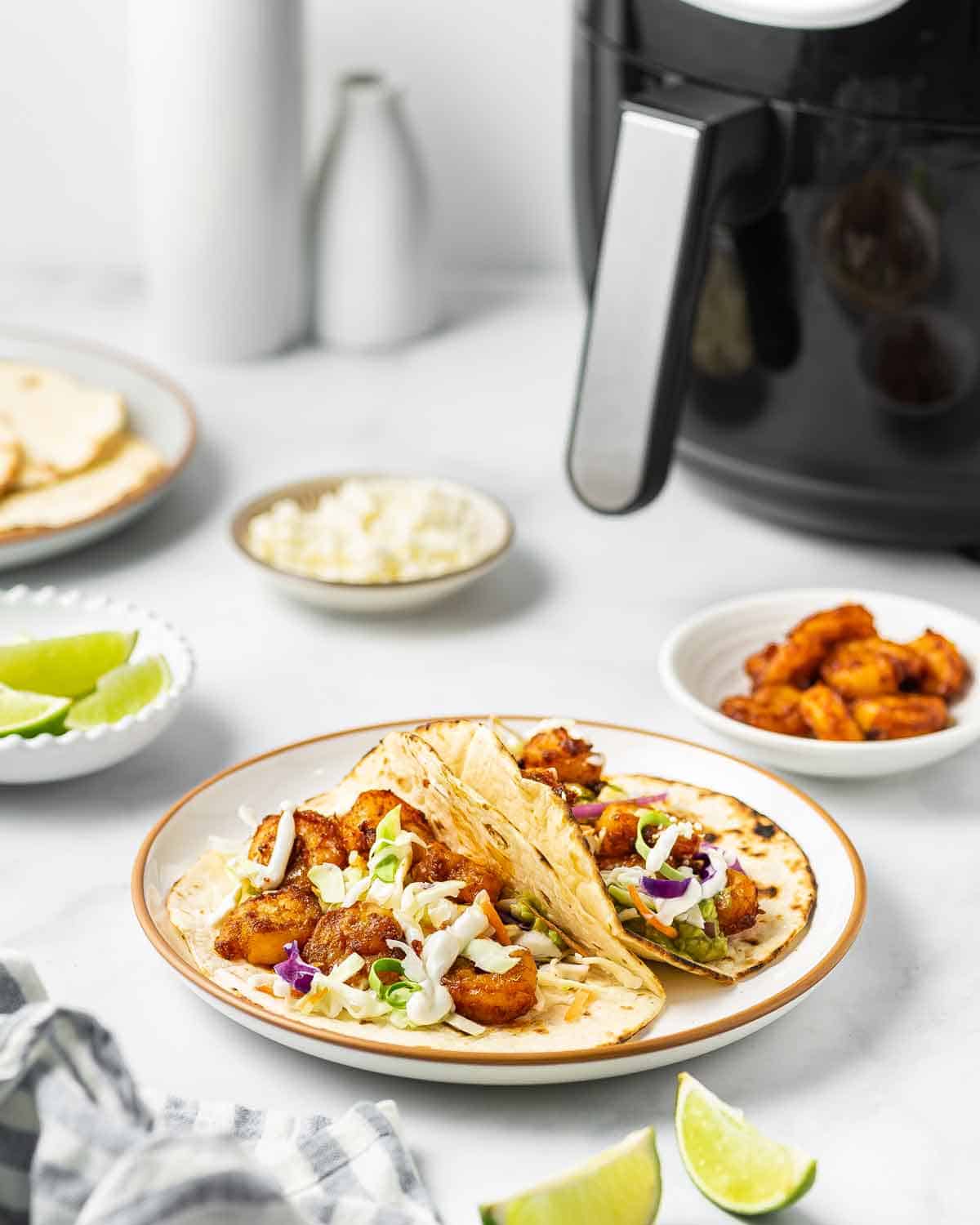 A plate with two shrimp tacos in front of an air fryer.