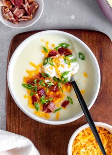 A bowl of potato soup with sour cream, shredded cheese, bacon, and green onions with a spoon inside.
