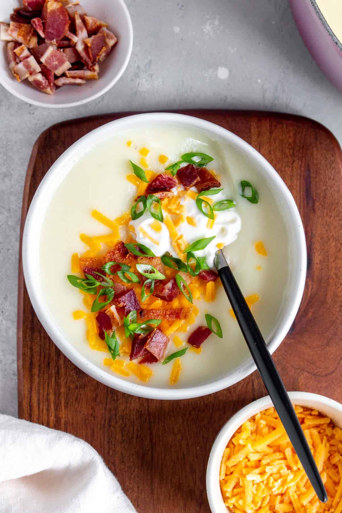 A bowl of potato soup with sour cream, shredded cheese, bacon, and green onions with a spoon inside.