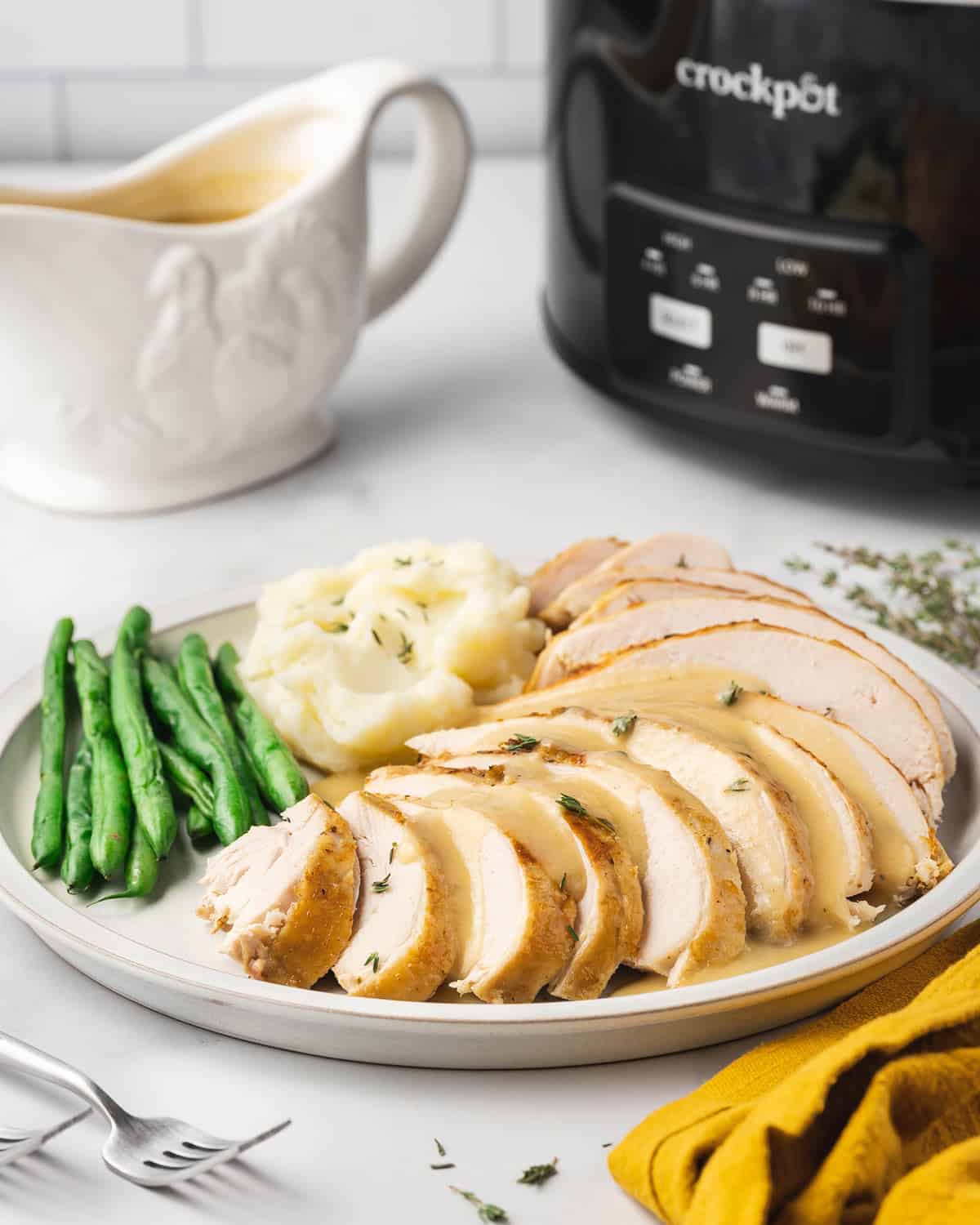 A plate of sliced turkey breast with green beans and mashed potatoes with a slow cooker in the background.