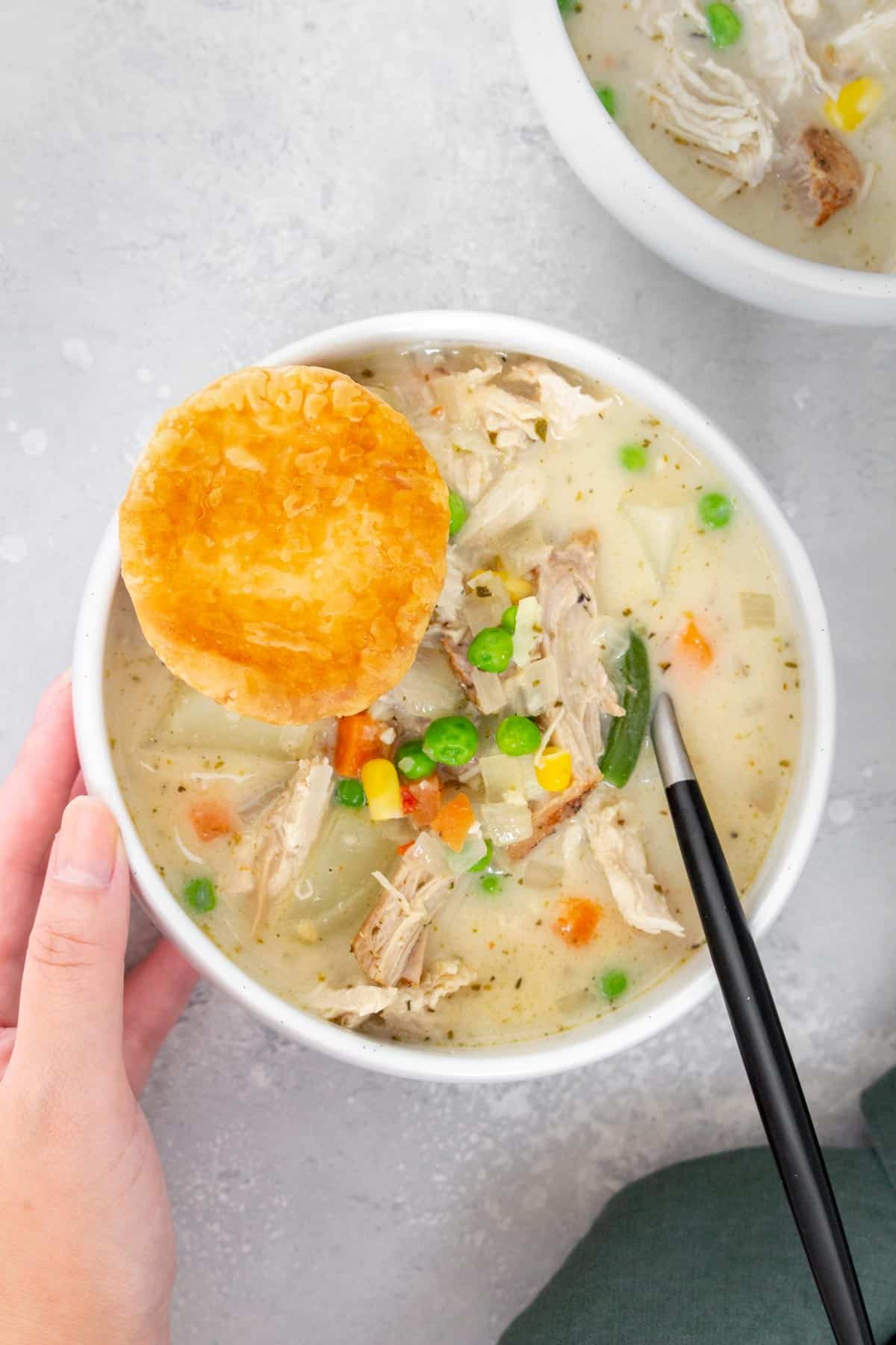 A bowl of turkey pot pie soup with a round puff pastry on the side with a hand on the side.