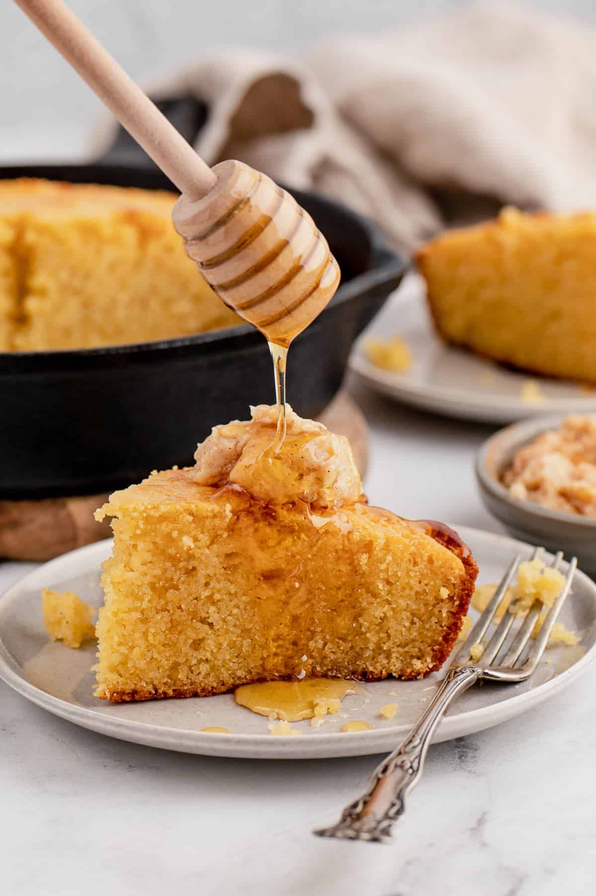 Honey on a stick drizzling over a slice of skillet cornbread on a plate with a fork.