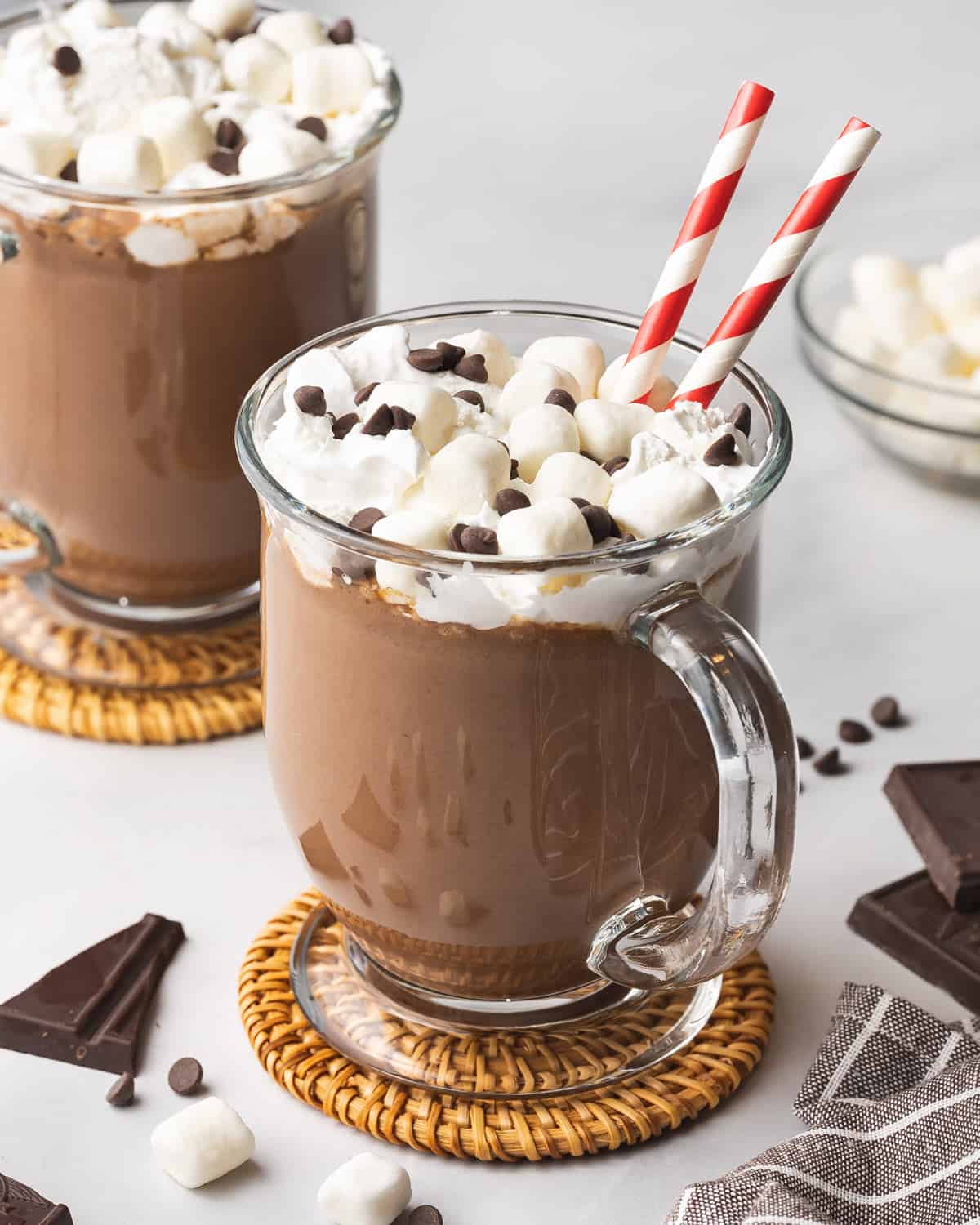 A glass mug of crockpot hot chocolate with whipped cream, mini marshmallows, mini chocolate chips and two straws.