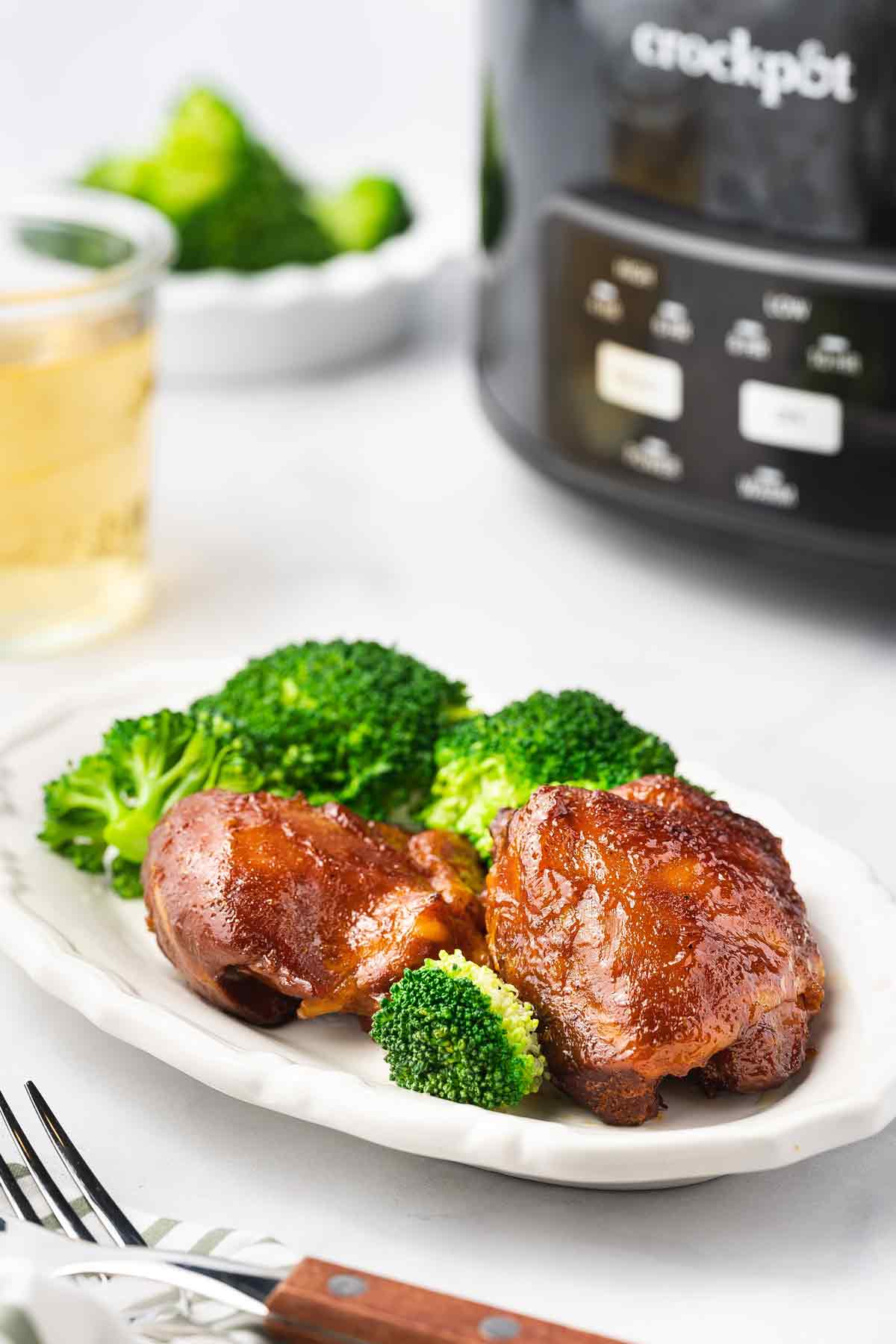 A plate of slow cooker bbq chicken thighs with broccoli with a crockpot in the background.
