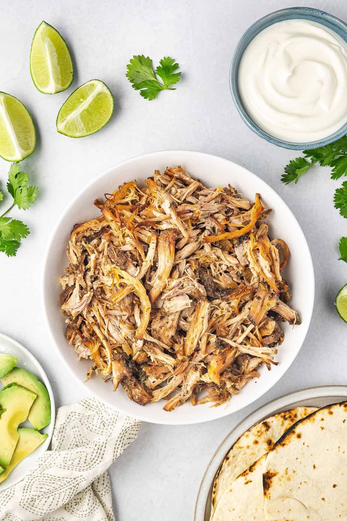 A bowl of shredded slow cooker carnitas with avocados, limes, sour cream, and toasted tortillas on the side.