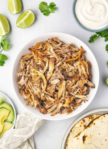 A bowl of shredded slow cooker carnitas with avocados, limes, sour cream, and toasted tortillas on the side.