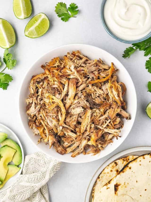 A bowl of shredded slow cooker carnitas with avocados, limes, sour cream, and toasted tortillas on the side.