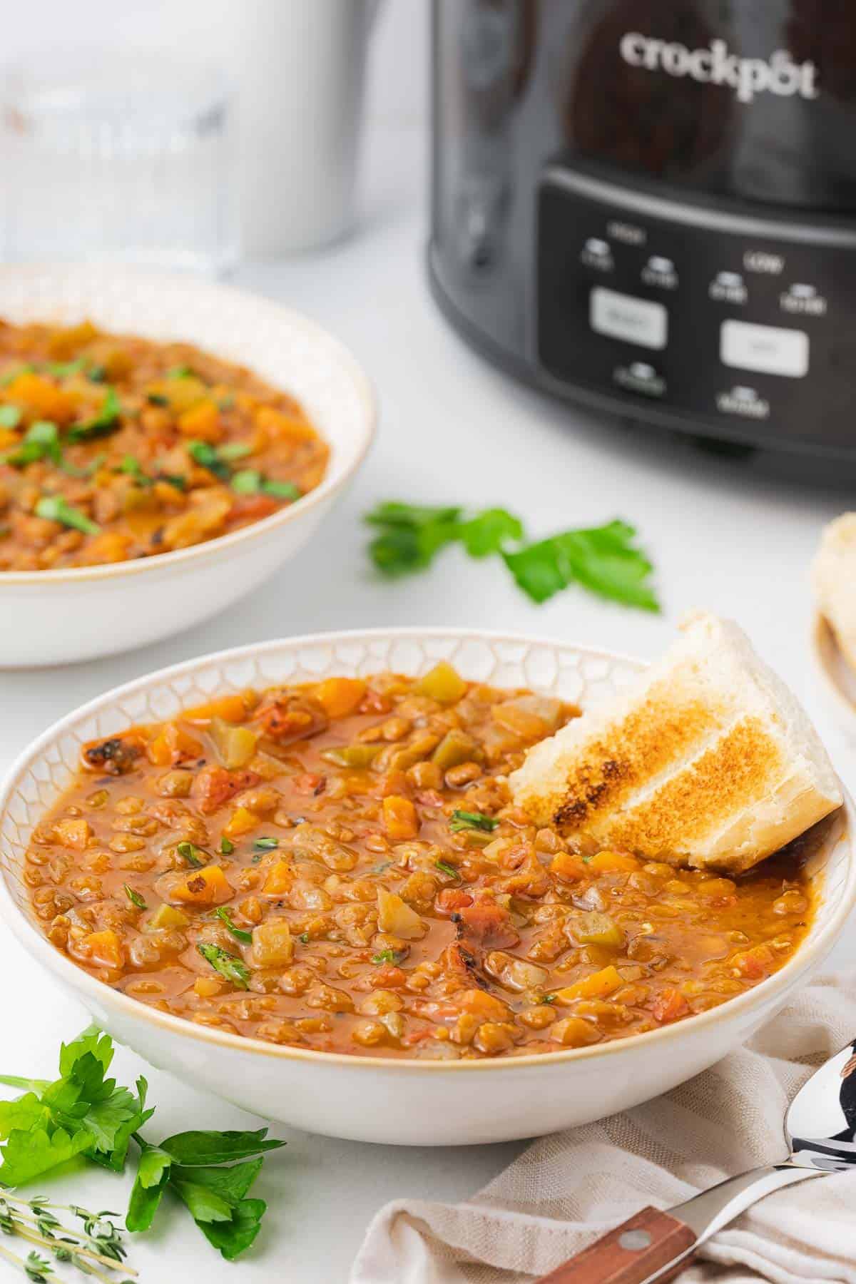 A bowl of slow cooker lentil soup with a piece of bread sticking out of it.