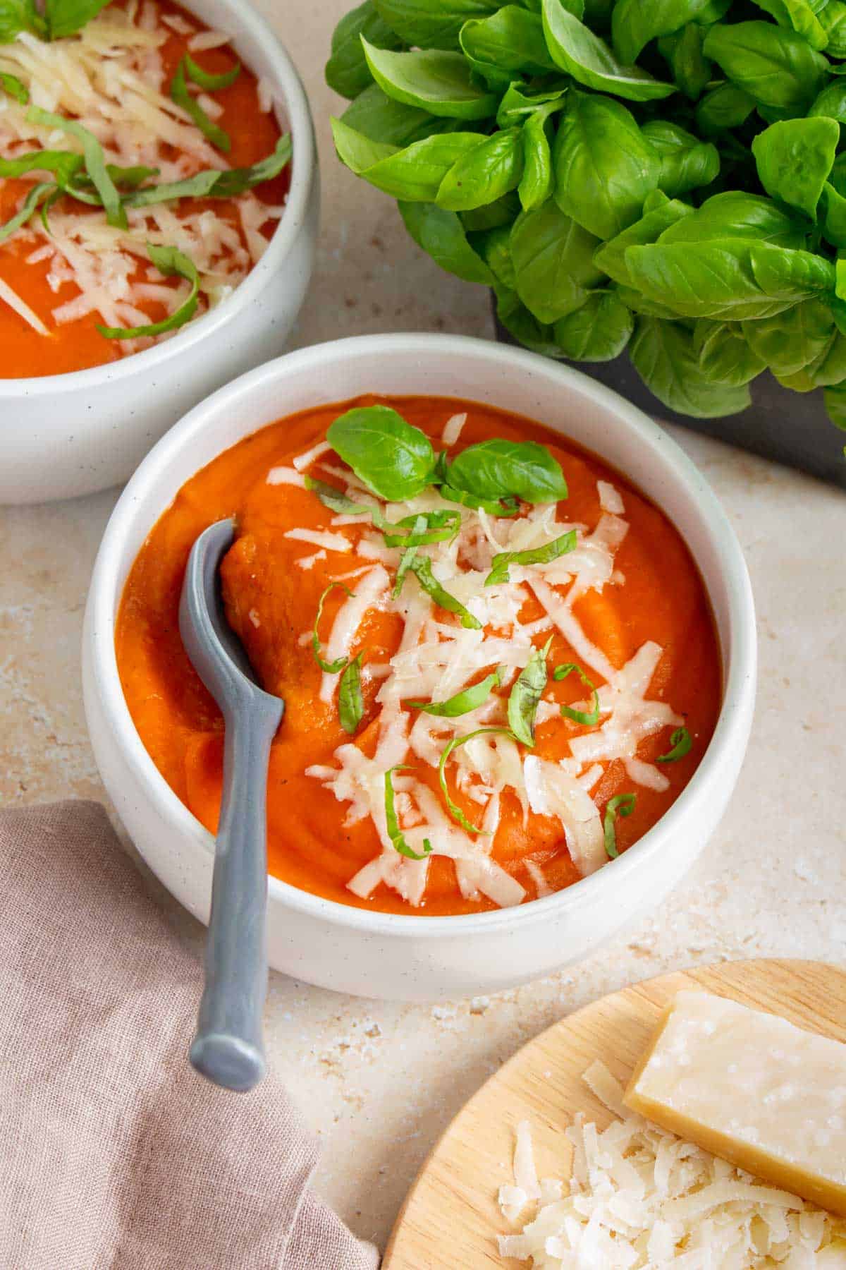 A bowl of potato tomato soup with parmesan and basil with a second bowl and a basil plant in the background. Shredded parmesan in the foreground.