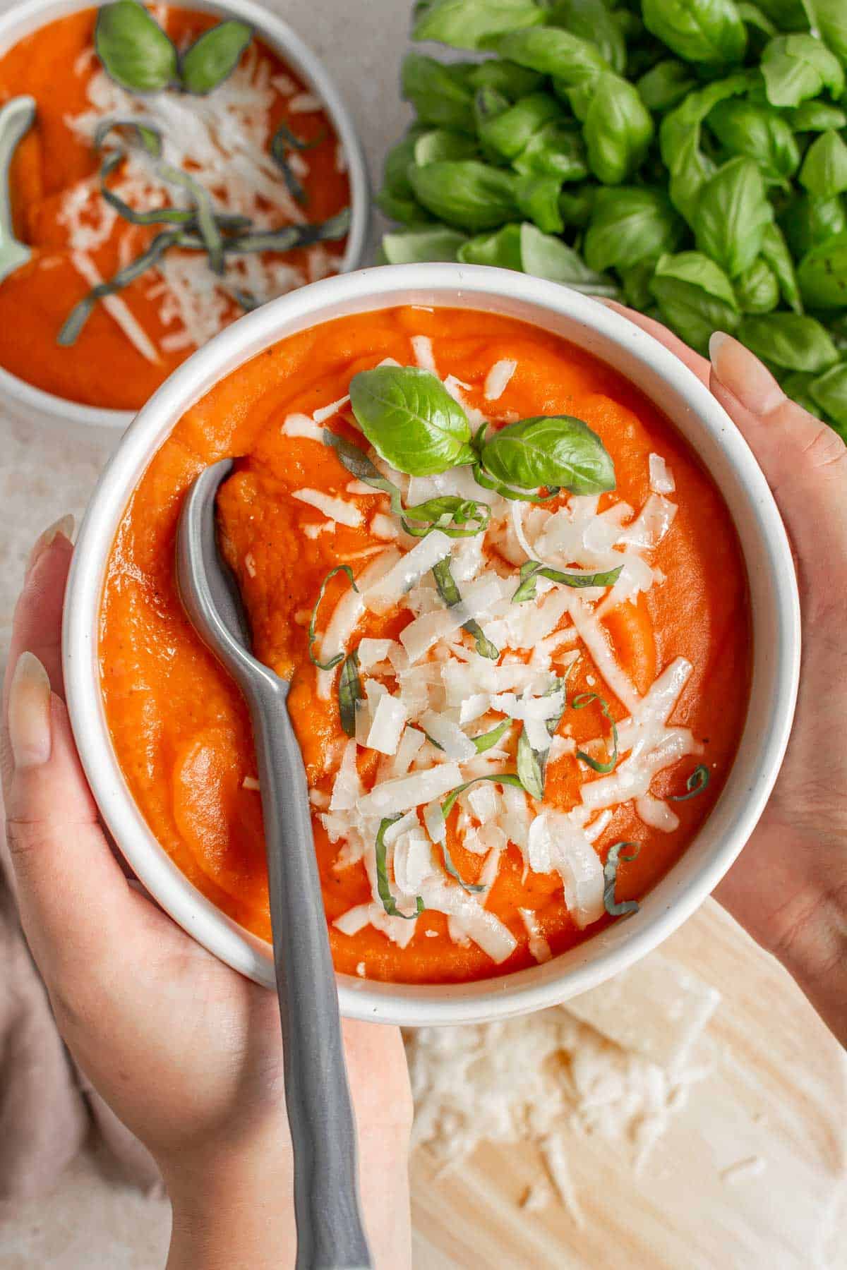 A hand holding up a bowl of potato tomato soup with a spoon inside with shredded parmesan and basil on top.