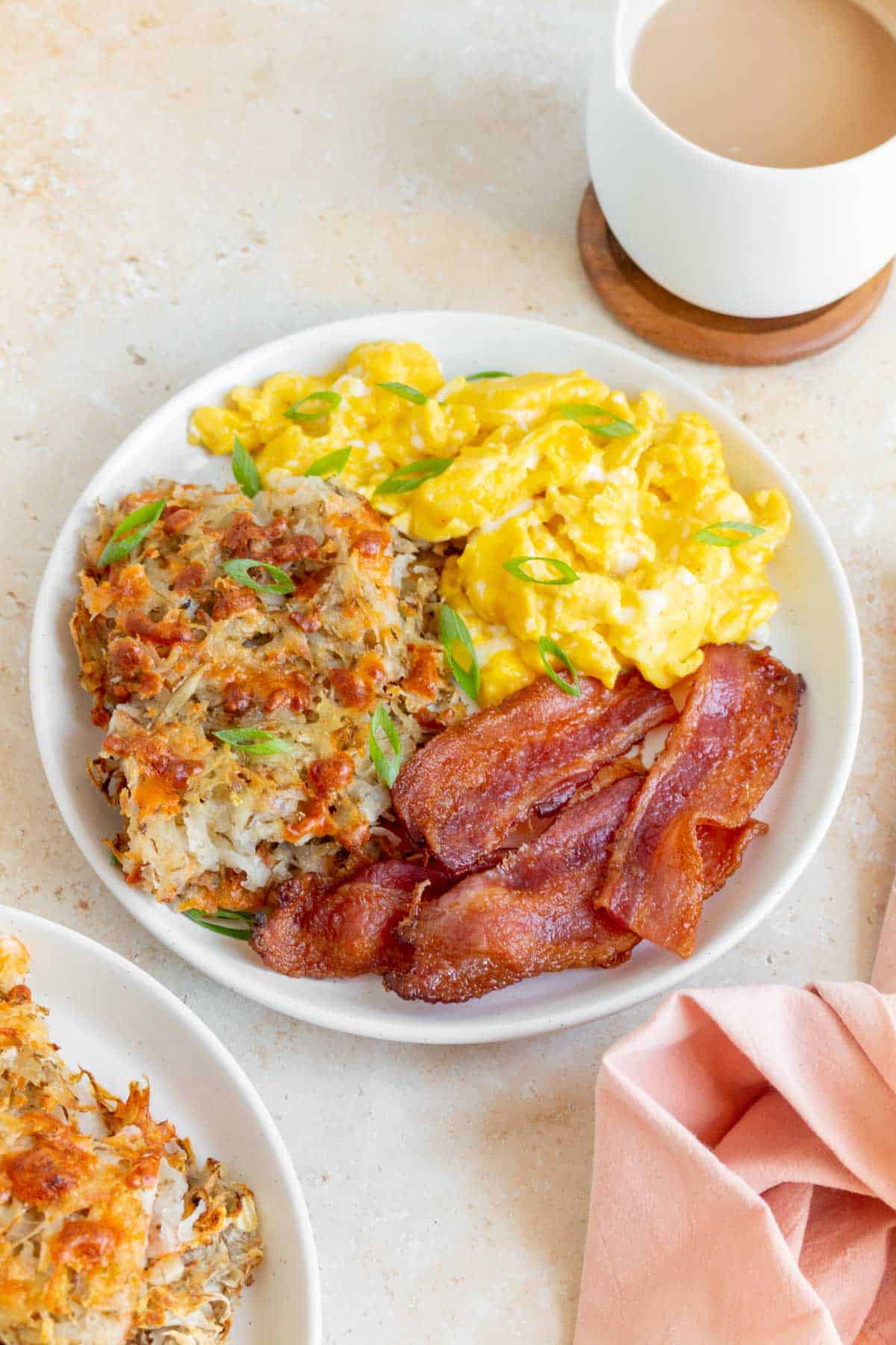 A plate with sheet pan hash browns, bacon, and scrambled eggs with a mug of coffee in the back.