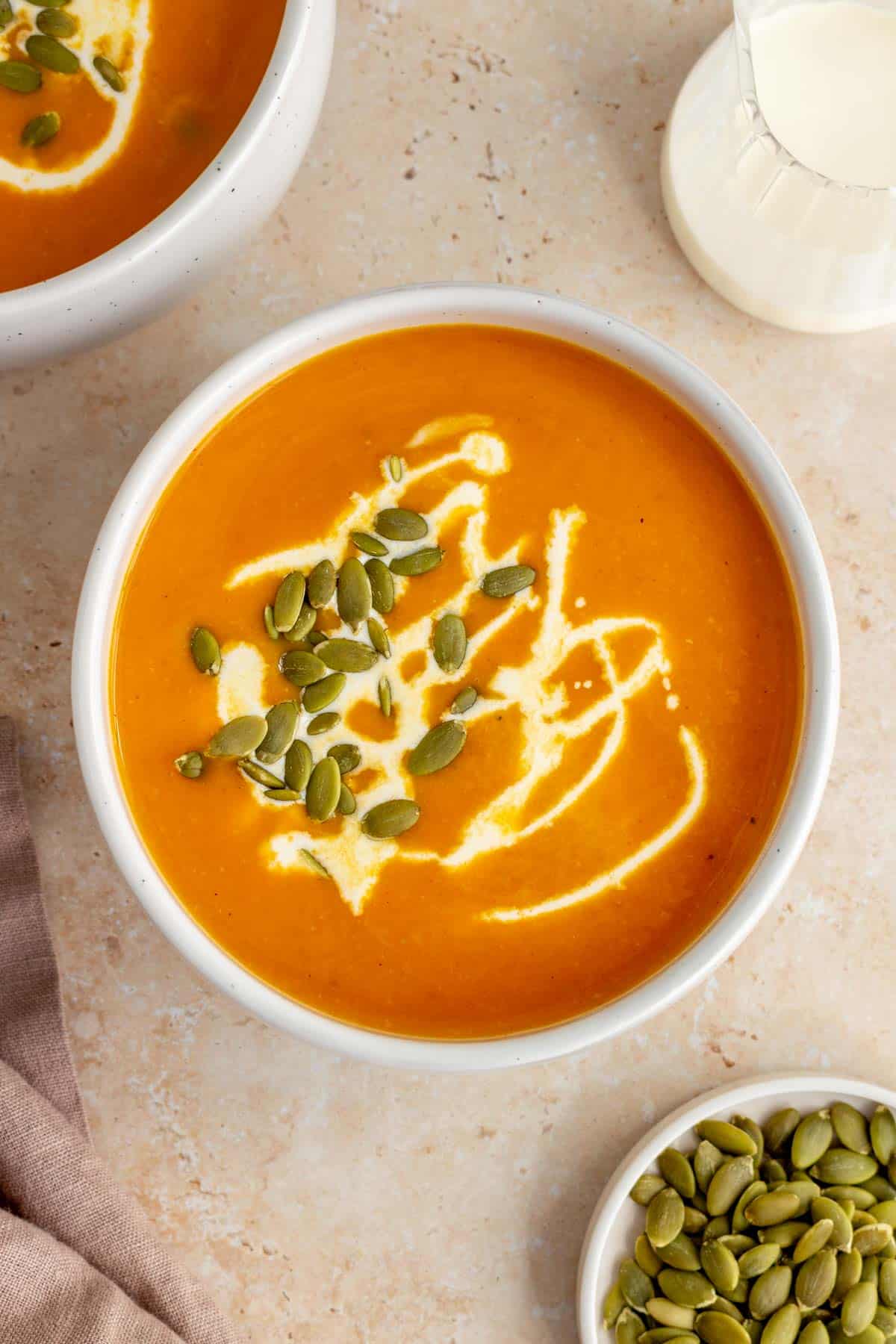Overhead view of a bowl of sweet potato and pumpkin soup garnished with coconut milk and pumpkin seeds. More coconut milk and pumpkins off to the side.