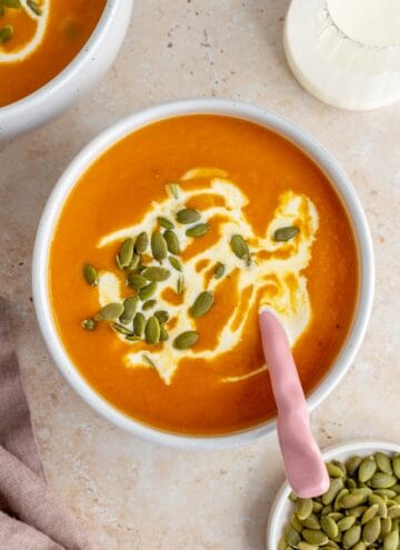 Overhead view of a bowl of sweet potato and pumpkin soup garnished with coconut milk and pumpkin seeds. A spoon in the soup.