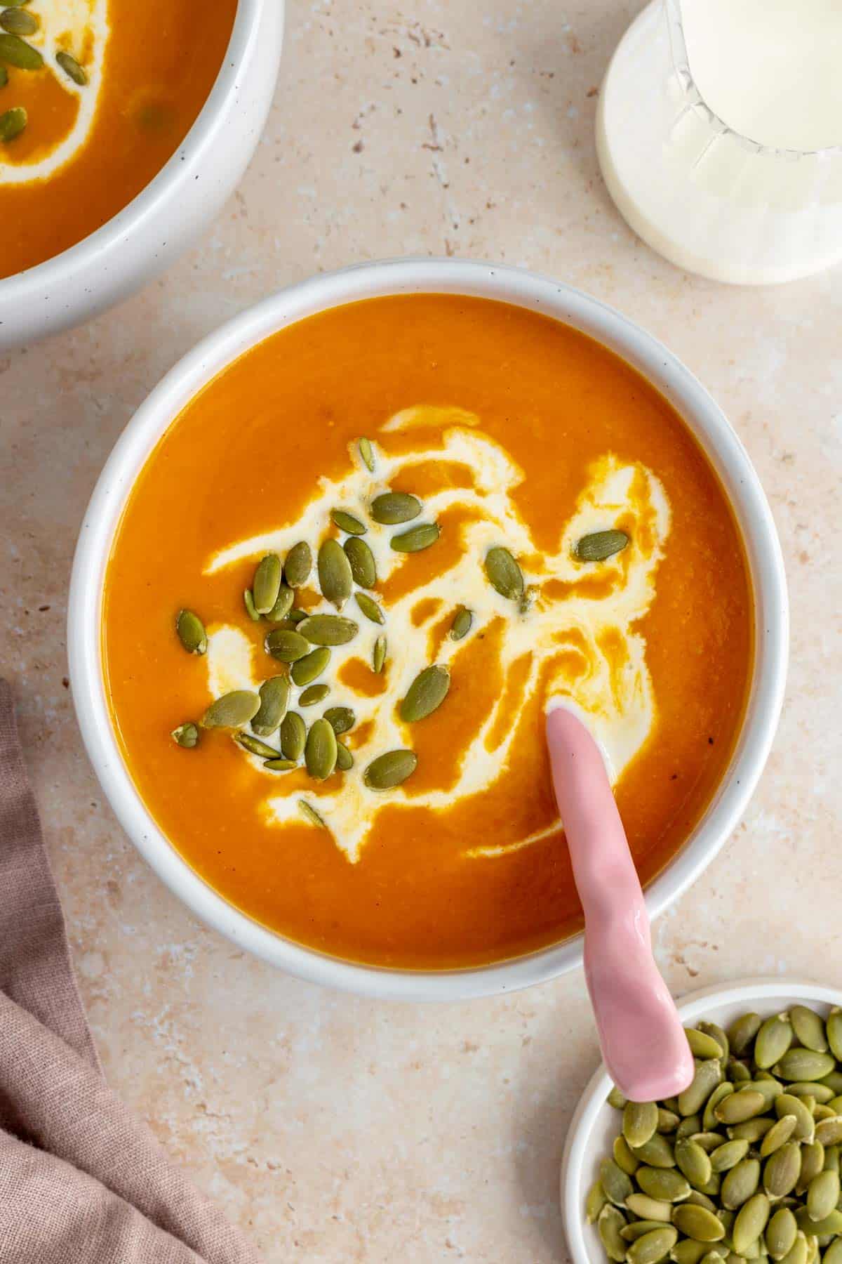 Overhead view of a bowl of sweet potato and pumpkin soup garnished with coconut milk and pumpkin seeds. A spoon in the soup.