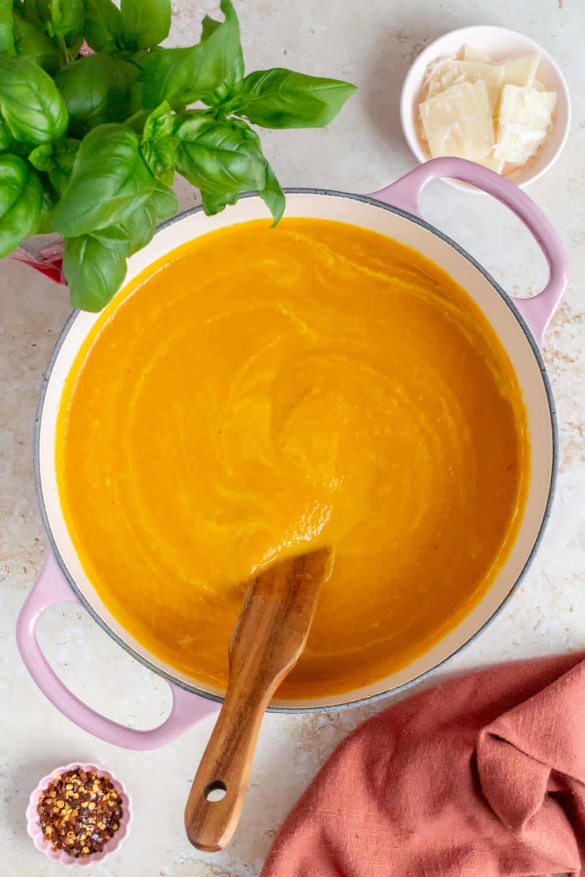 Overhead view of a pot of carrot pumpkin soup surrounded by fresh basil, parmesan, and red pepper flakes.