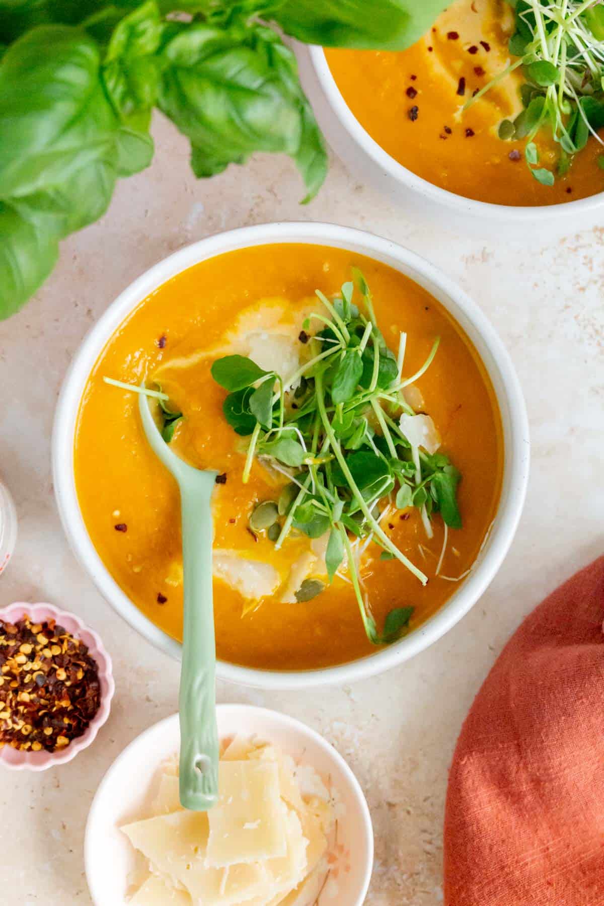 Overhead view of a bowl of carrot pumpkin soup with a green spoon with some microgreens, basil, and parmesan on top with more off to the side.