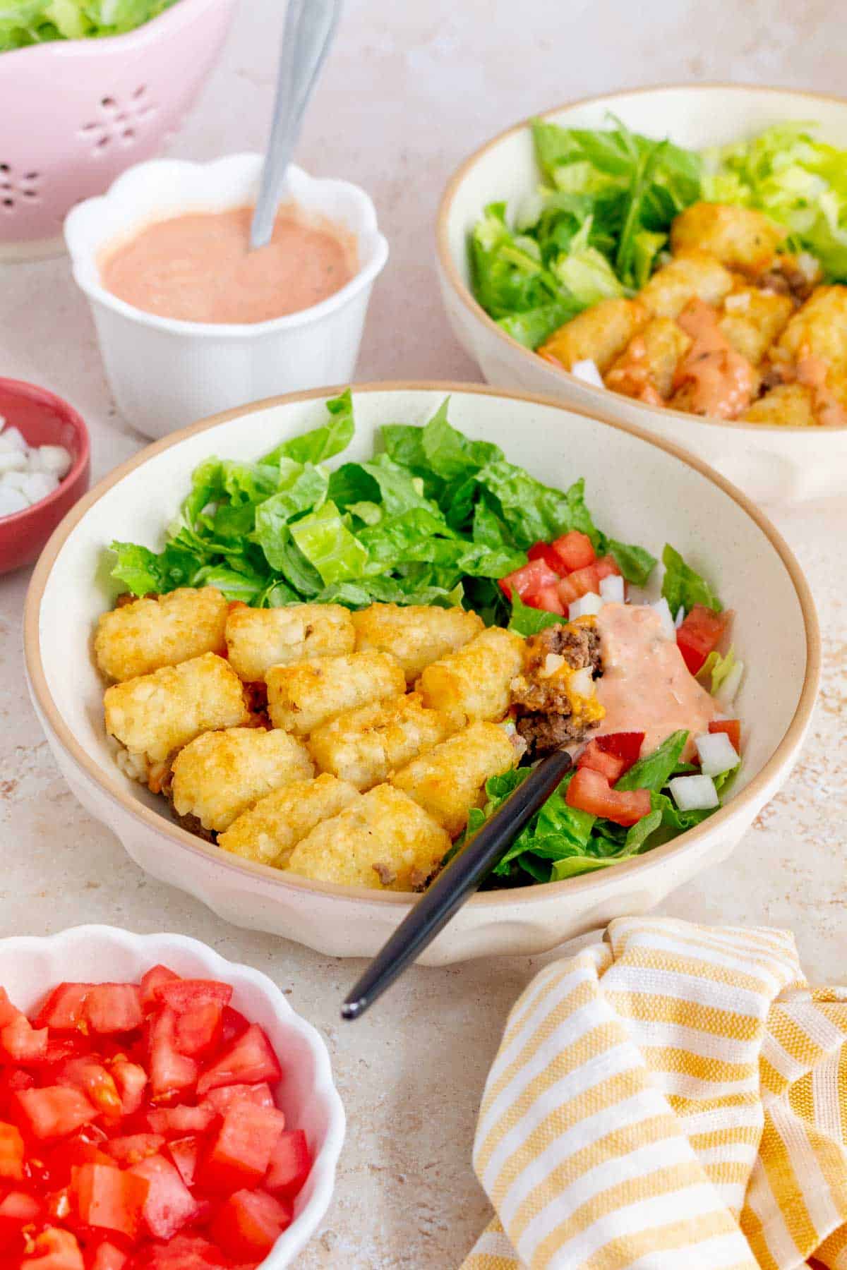 A bowl of big mac casserole with tater tots with lettuce, tomatoes, onions, and big mac sauce. A second bowl in the back and toppings in bowls scattered around.