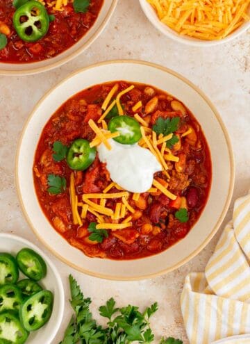 A bowl of Dutch oven chili topped with sour cream, shredded chili, jalapeno, and cilantro. More toppings in bowls on the side.