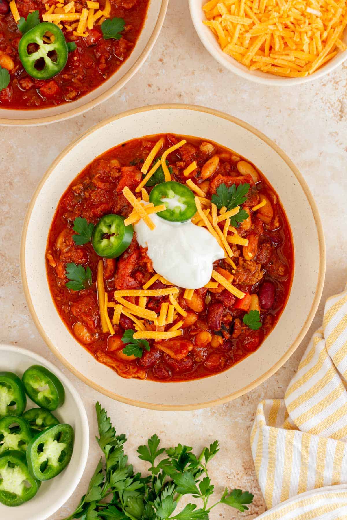 A bowl of Dutch oven chili topped with sour cream, shredded chili, jalapeno, and cilantro. More toppings in bowls on the side.