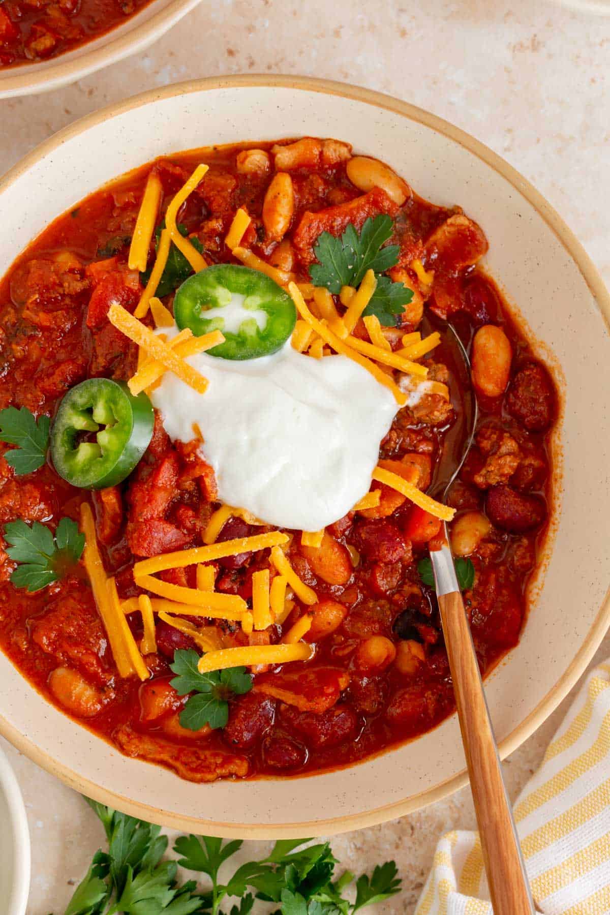 A close up of a bowl of Dutch oven chili topped with sour cream, shredded chili, jalapeno, and cilantro with a spoon inside.