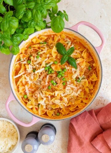 Overhead view of a pink Dutch oven of ground pork pasta topped with parmesan and basil leaves.