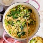 A pink dutch oven with cheesy broccoli orzo. A wooden spoon inside.