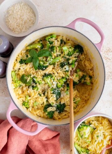 A pink dutch oven with cheesy broccoli orzo. A wooden spoon inside.