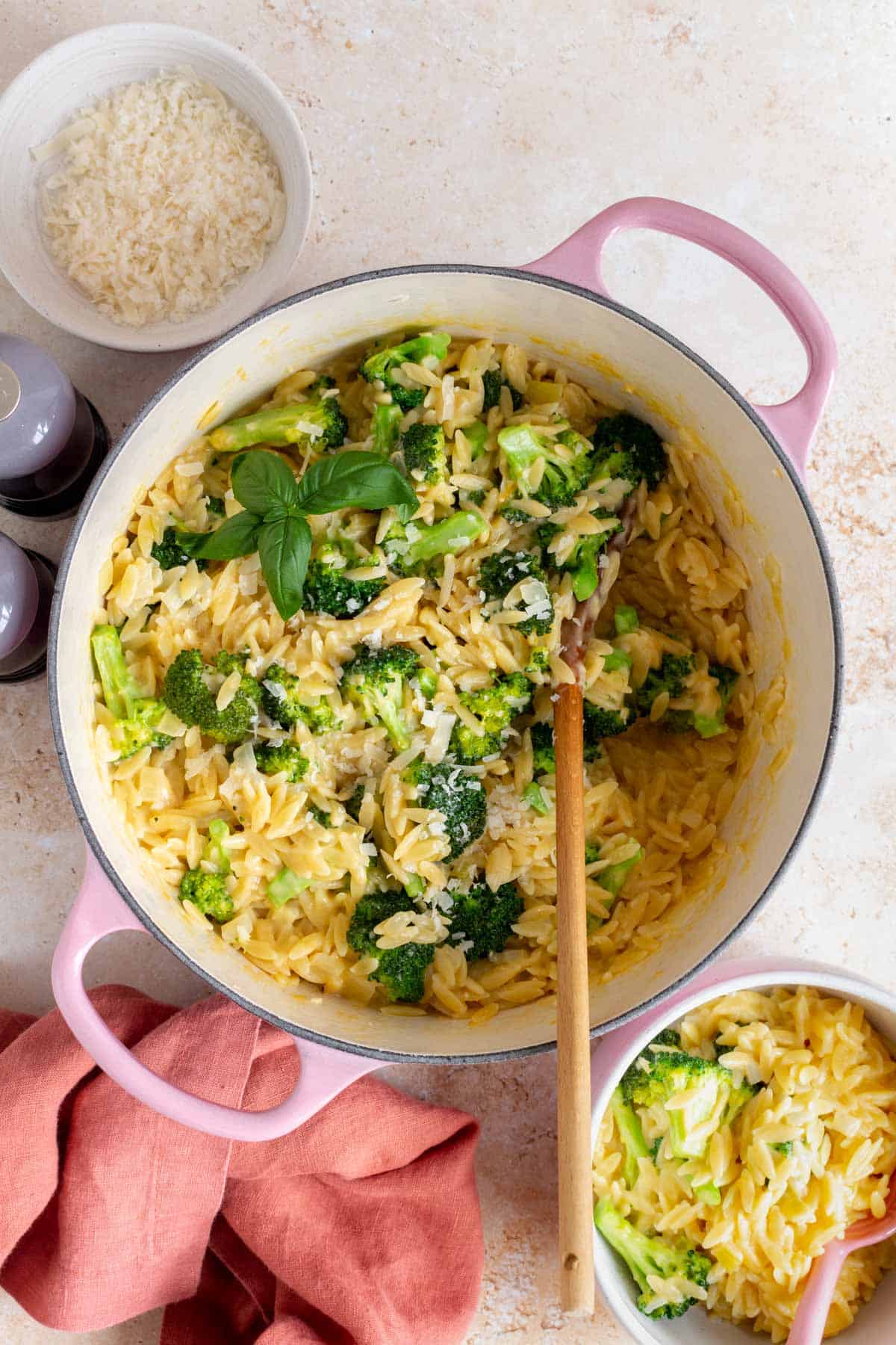 A pink dutch oven with cheesy broccoli orzo. A wooden spoon inside.