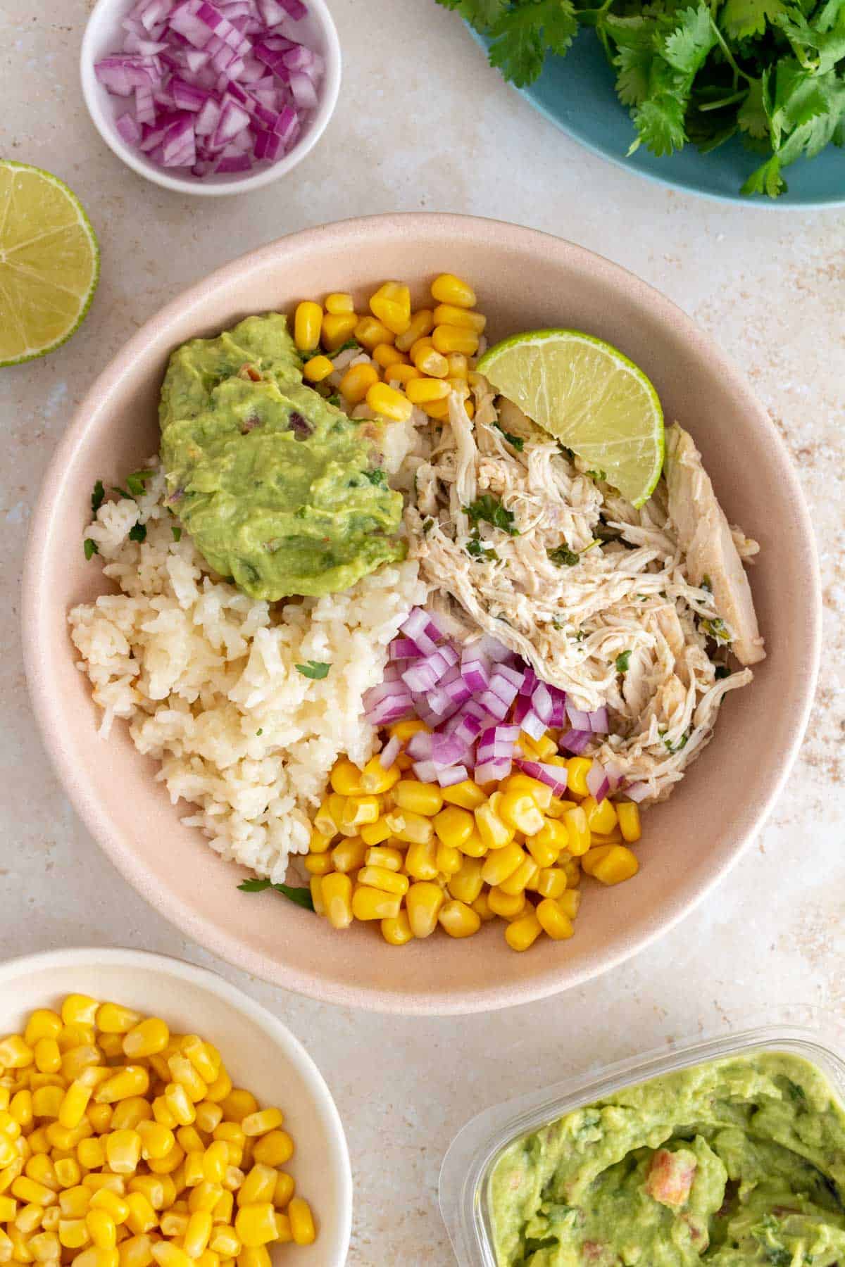 A bowl of rice, corn, guacamole, red onions, lime wedge, and shredded slow cooker cilantro lime chicken.