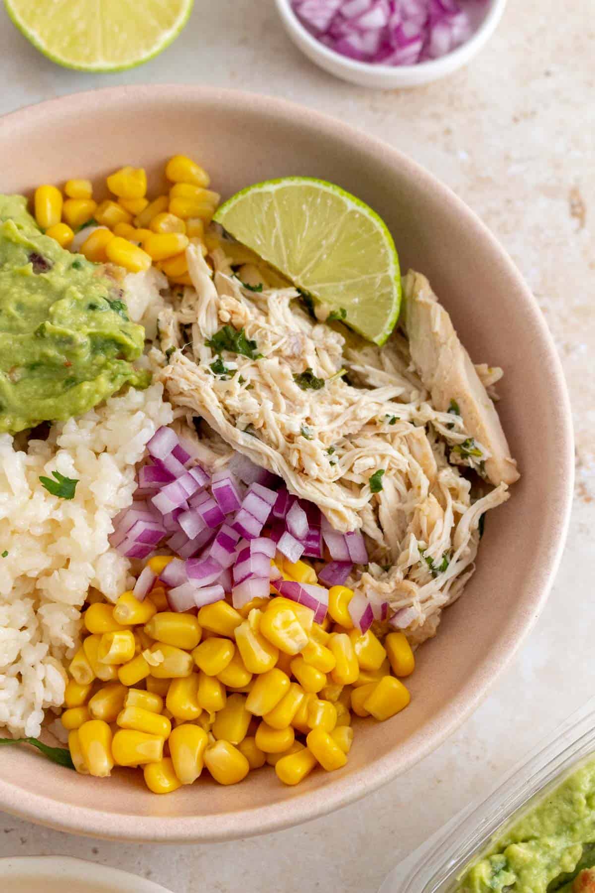 A close up view of shredded slow cooker cilantro lime chicken in a bowl with rice, corn, red onions, guacamole, and a lime wedge.