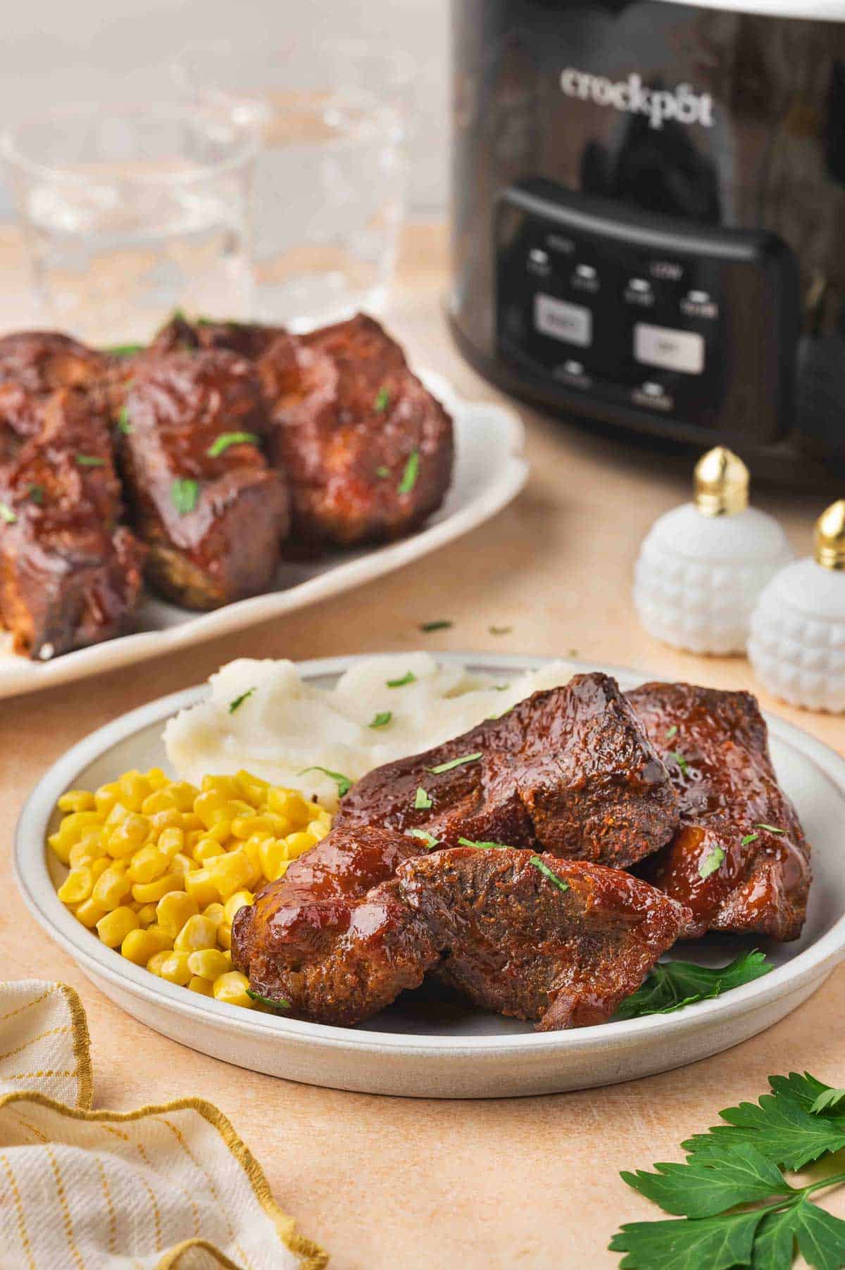 A plate of slow cooker country style ribs with corn and mashed potatoes with a slow cooker in the background and more ribs.