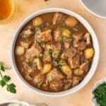 Overhead view of a bowl of slow cooker beef tips and gravy surrounded by a bowl of mashed potatoes, two drinks, and fresh herbs.