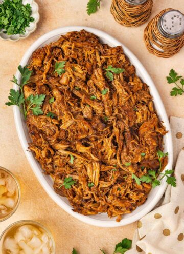 Overhead view of a platter of slow cooker gochujang pulled pork garnished with parsley. Salt and pepper shakers on the side along with some iced drinks.
