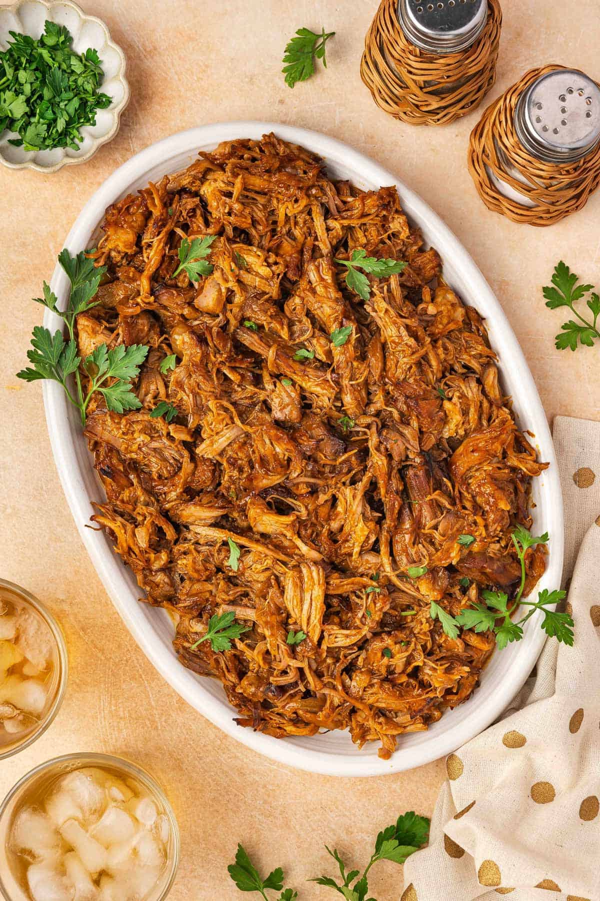 Overhead view of a platter of slow cooker gochujang pulled pork garnished with parsley. Salt and pepper shakers on the side along with some iced drinks.