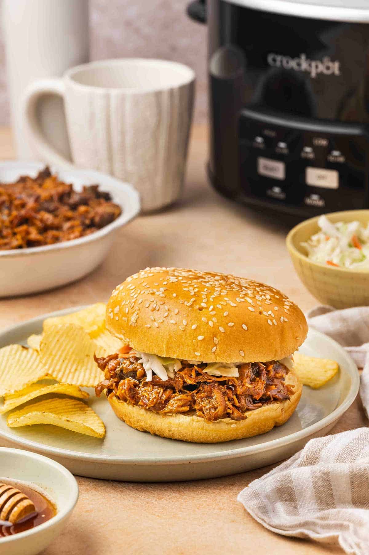 A plate with shredded slow cooker hot honey chicken inbetween burger buns with some chips on the side and a slow cooker in the background.