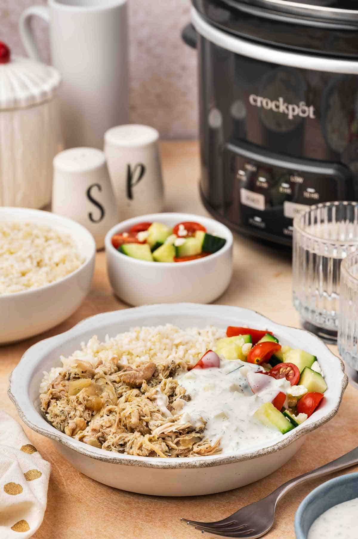A bowl of shredded slow cooker Greek chicken along with some rice, cucumber, tomatoes, and tzatziki sauce. A slow cooker in the background.