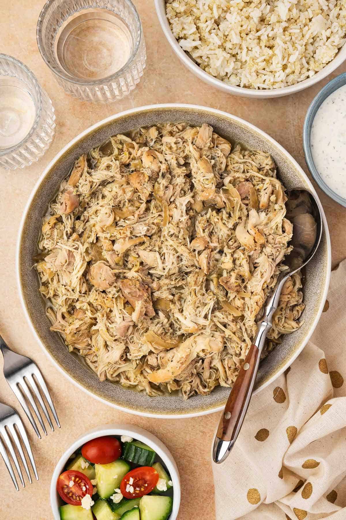 Overhead view of a bowl of shredded slow cooker Greek chicken along with a spoon. Some rice, salad, forks, drinks, and a napkin on the side.