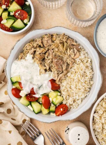 A bowl of shredded slow cooker Greek chicken along with some rice, cucumber, tomatoes, and tzatziki sauce.