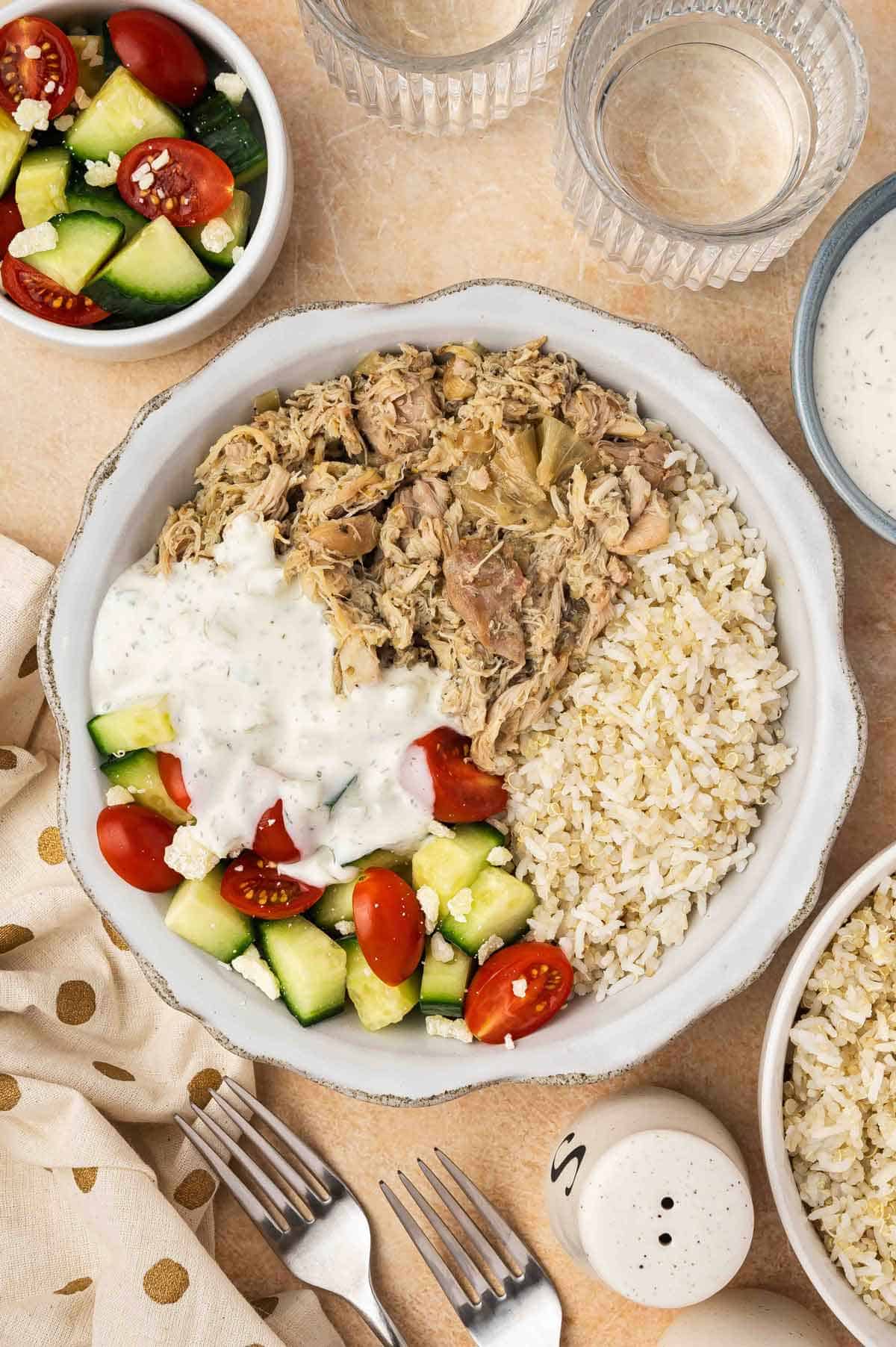 A bowl of shredded slow cooker Greek chicken along with some rice, cucumber, tomatoes, and tzatziki sauce.