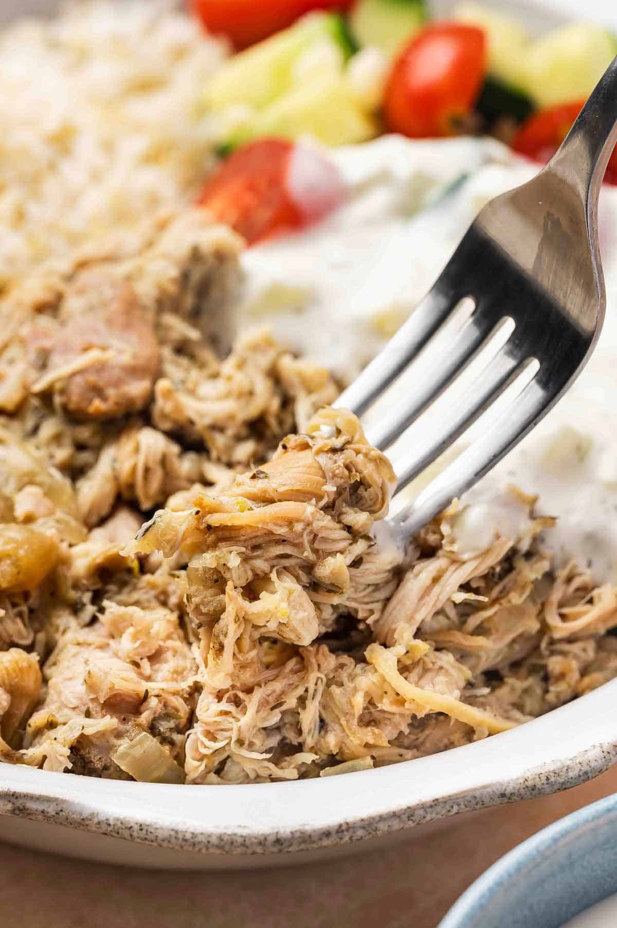 A fork lifting up some shredded slow cooker Greek chicken from a bowl.