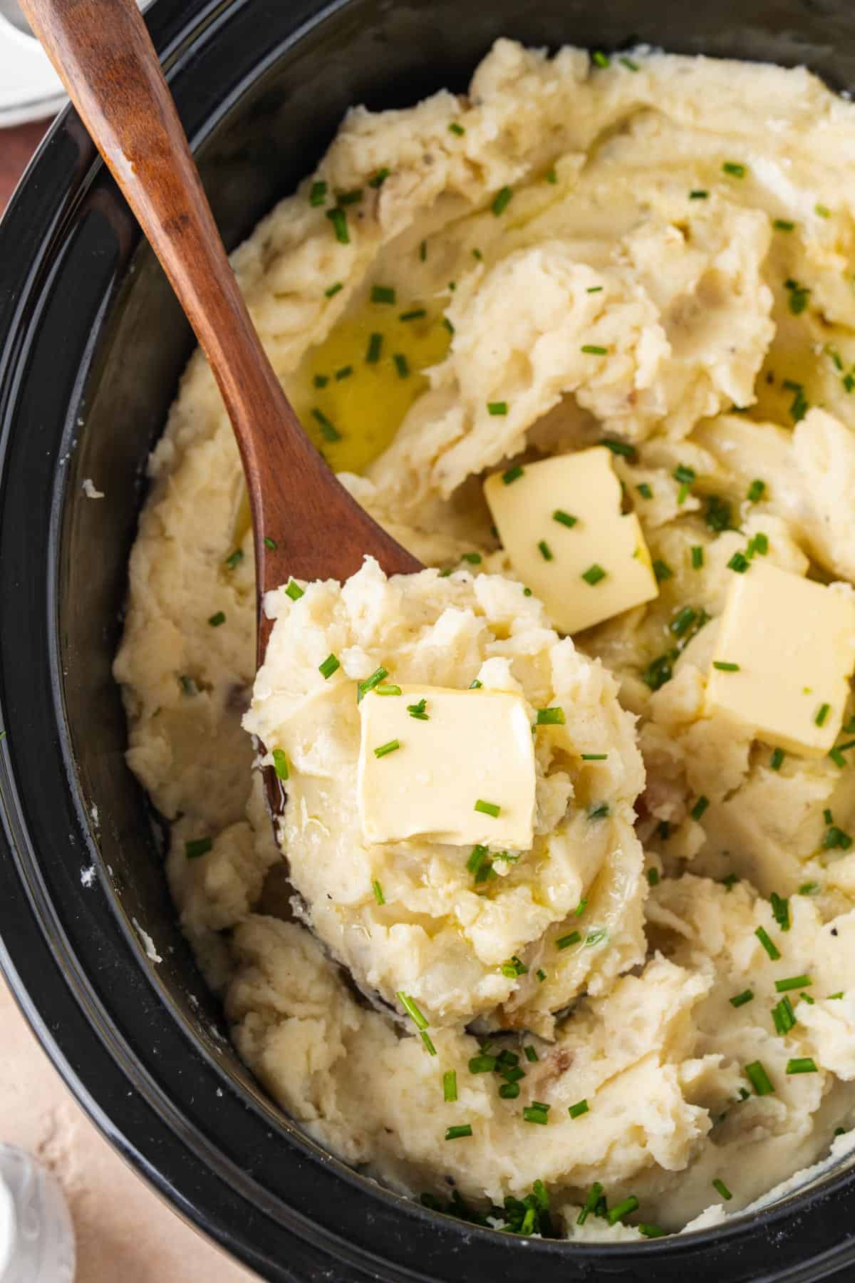 A close up view of a spoonful of mashed potatoes with garlic and sour cream in a slow cooker topped with butter and chives.