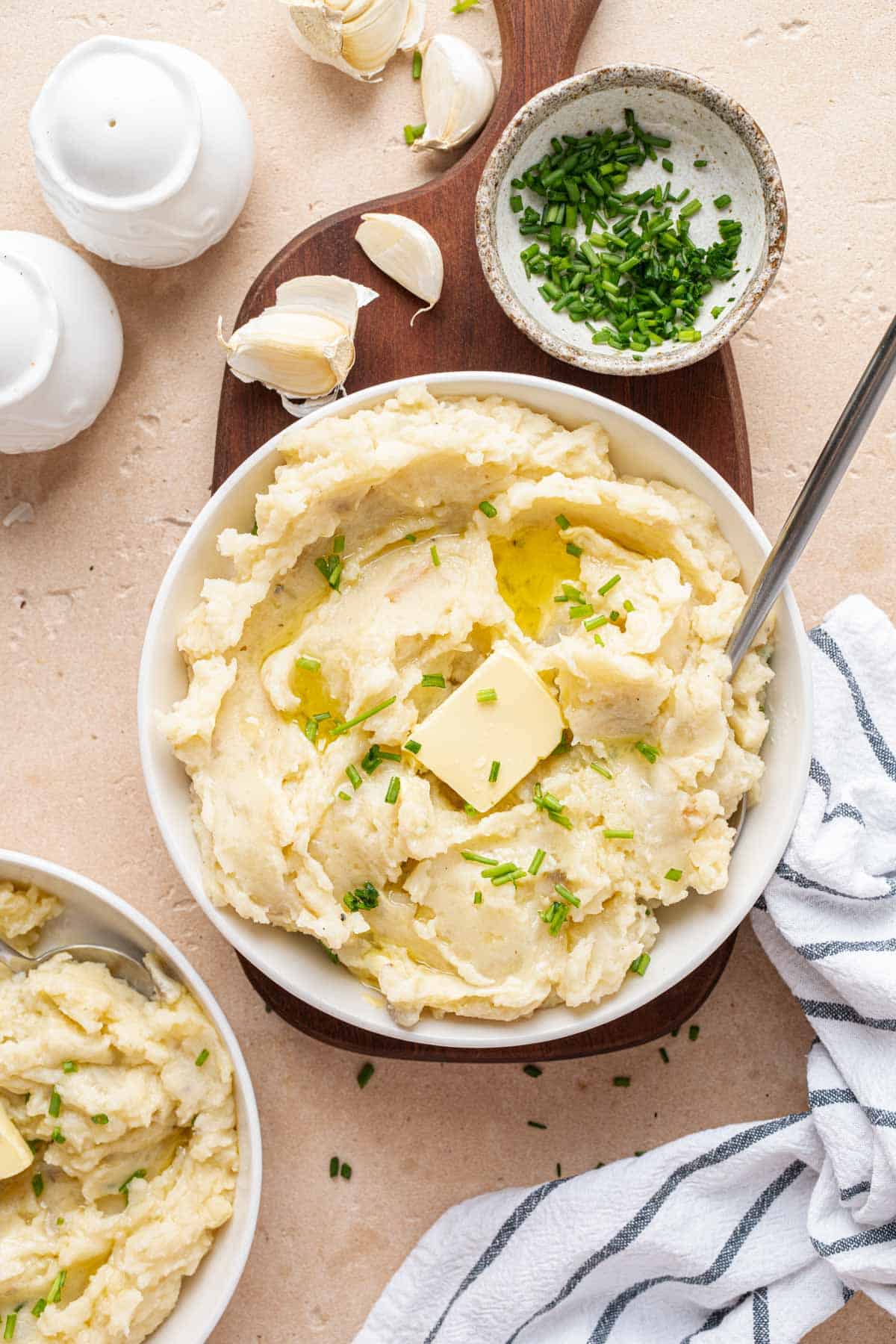 A plate with mashed potatoes topped with butter and chives. A bowl of chopped chives and some garlic cloves on the side.